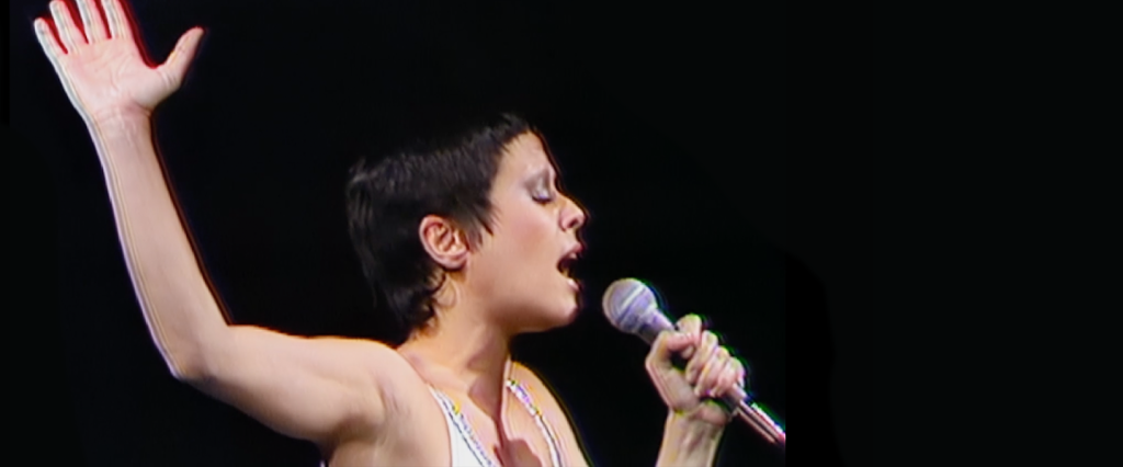 A cantora Elis Regina, segurando um microfone, de olhos fechados e com uma das mãos para cima