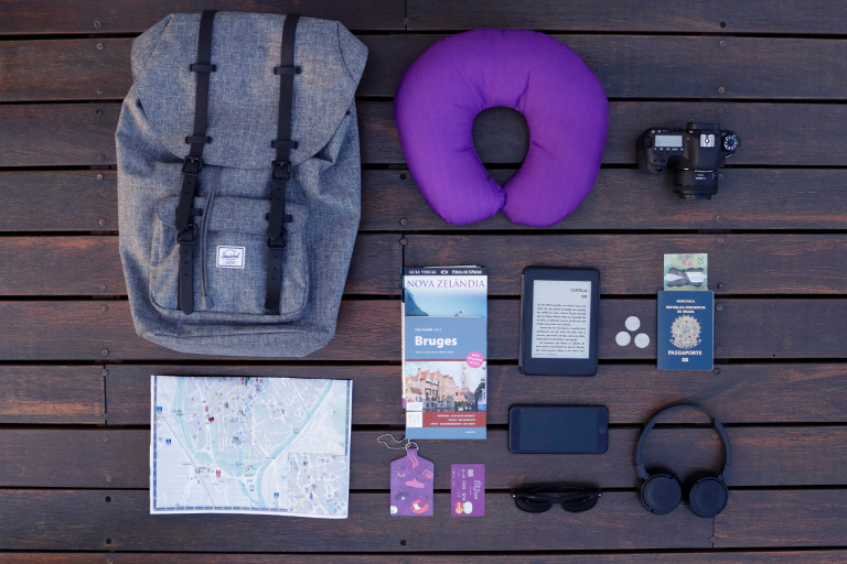 Diversos itens de viagem vistos de cima: mochila, mapa, almofada de pescoço, máquina fotográfica, passaporte e cartão Nubank