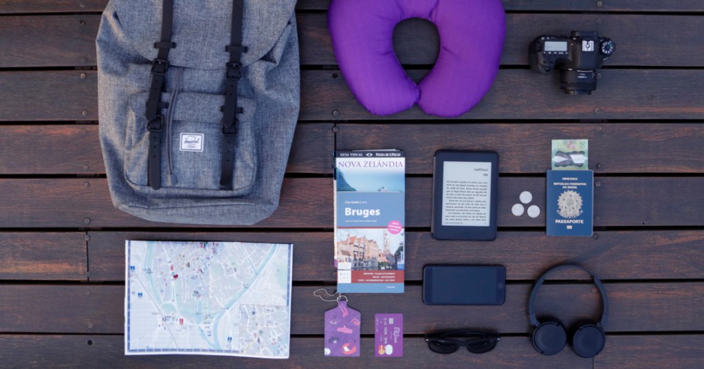 Diversos itens de viagem vistos de cima: mochila, mapa, almofada de pescoço, máquina fotográfica, passaporte e cartão Nubank