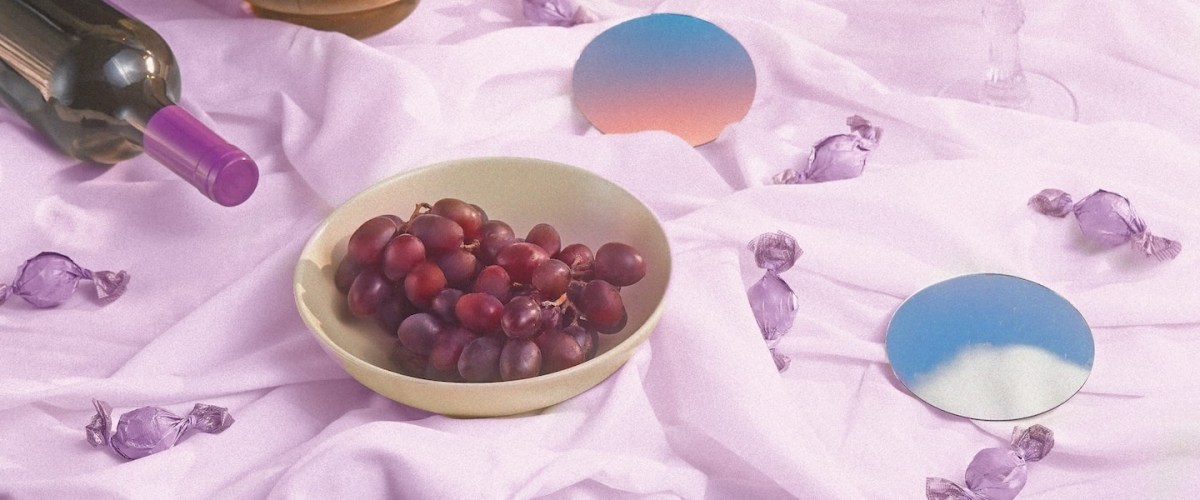 Imagem de uma mesa forrada com um tecido roxo. Sobre ela estão: duas taças com vinho, uma tigela com uvas e outra com pipoca e uma garrafa de vinho. Há também bombons espalhados pela mesa.