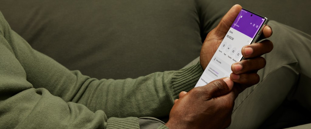 O que é o Nubank? Imagem mostra as mãos de um homem negro vestindo uma camiseta de manga comprida cor verde musgo e segurando um celular na tela inicial do app do Nubank enquanto está sentado em um sofá também na cor verde musgo.