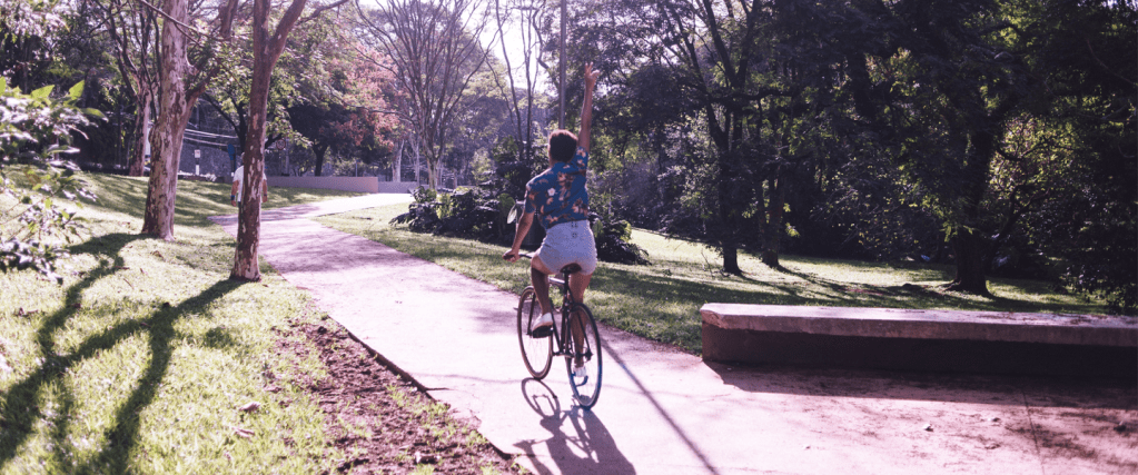 Jovem, de costas, andando de bicicleta com uma das mãos para cima. O dia está ensolarado e a bicicleta está em um parque com árvores