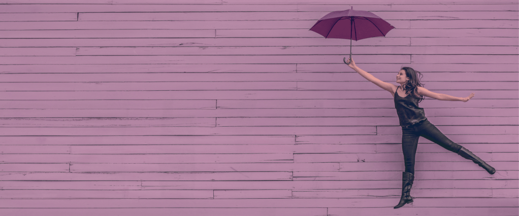 Jovem pulando segurando um guarda-chuva roxo em frente a uma parede lilás