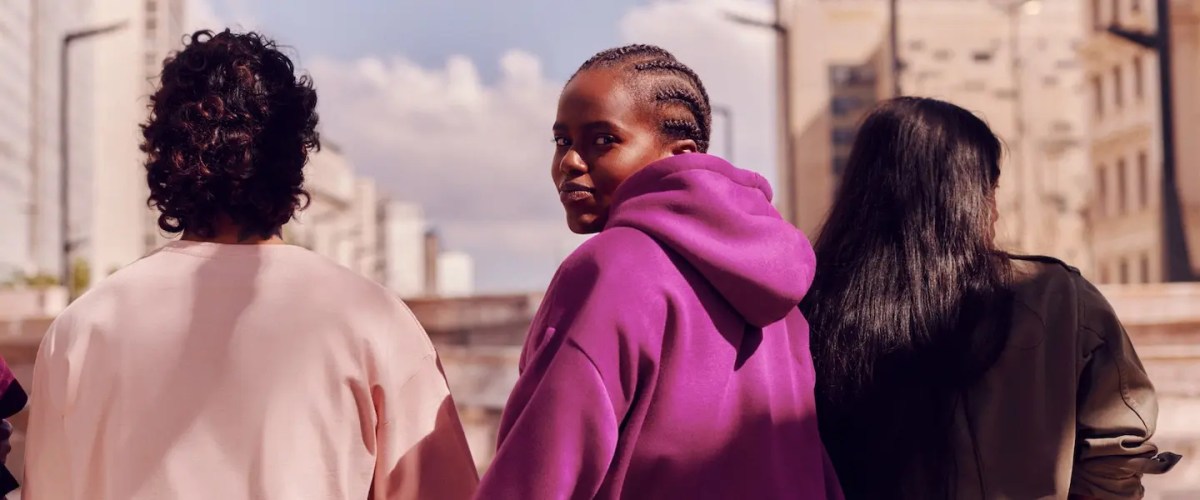 O que é o Nubank: imagem de três pessoas sentadas na direção oposta à câmera, de frente para prédios. A pessoa do meio, uma mulher com tranças no cabelo e um moletom rosa, se vira para a câmera e olha por cima do ombro.