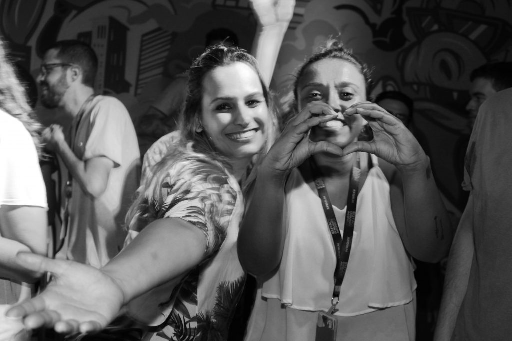 Duas jovens em uma festa, sorrindo para a câmera. Uma delas está fazendo um coração com as mãos. A foto é em preto e branco