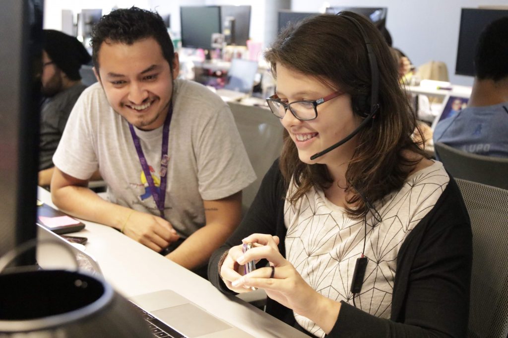 Um homem e uma mulher sentados em frente ao computador, sorrindo. Ela está usando um fone com microfone embutido