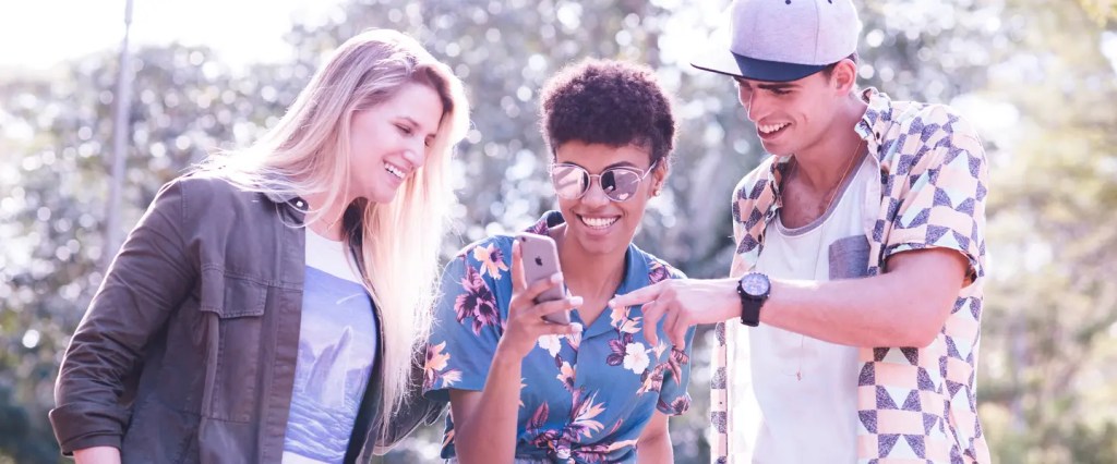 Três pessoas, duas mocas e um rapaz, de pé, olhando para o telefone na mão da jovem do meio.