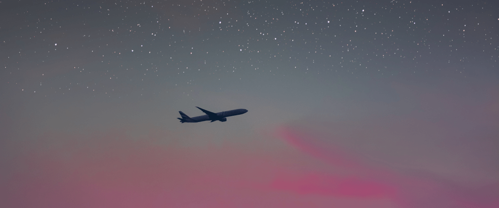 A foto mostra um avisão voando em um céu ao anoitecer. O céu está rosa na linha do horizonte e cheio de estrelas mais acima