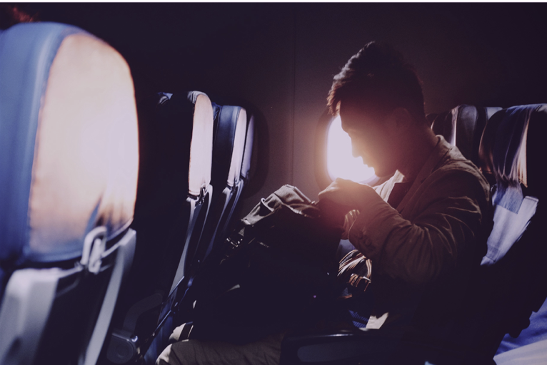 Foto mostra passageiro sentado na janelinha do avião, mexendo em uma mochila, e com o sol alaranjado visto pelo vidro