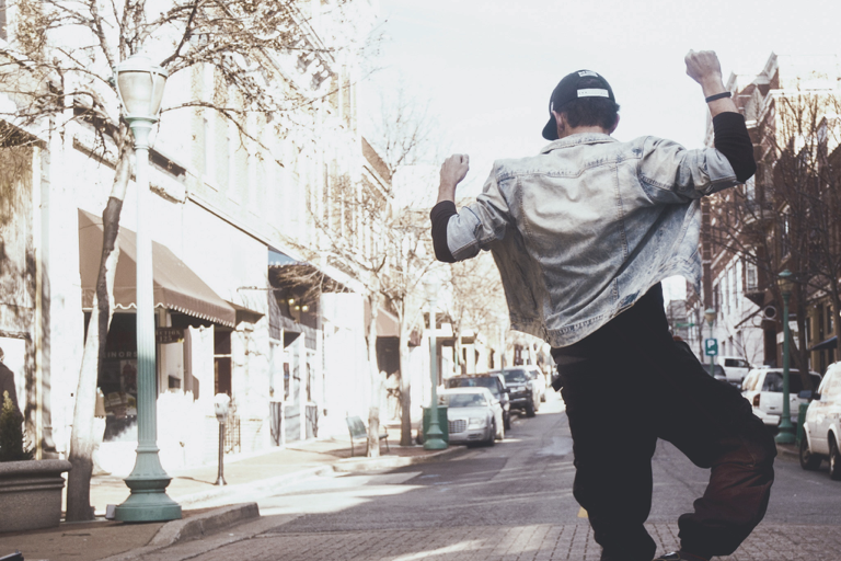 Jovem de bone, jaqueta jeans e calça preta visto de costas pulando no meio da rua com os braços para cima