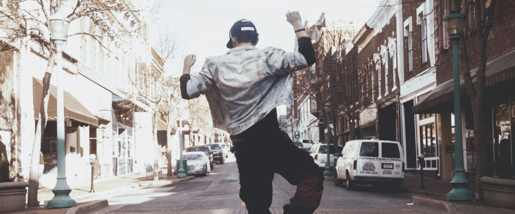 Jovem de boné, jaqueta jeans e calça preta visto de costas pulando no meio da rua com os braços para cima