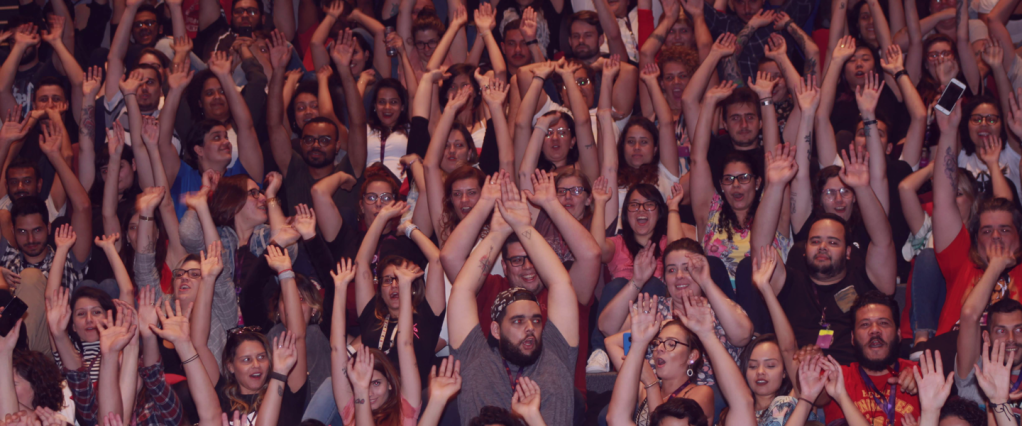 grupo de pessoas em um auditório lotado, todas com as mãos para cima