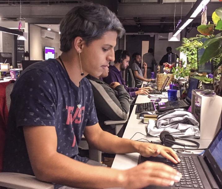 Foto mostra bancada de trabalho, toda decorada com plantas, e um membro da equipe de atendimento do Nubank digitando em um computador