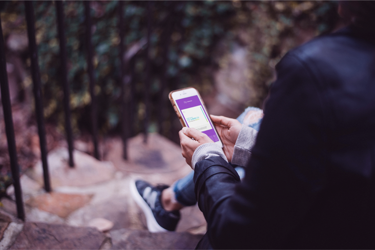 Pessoa sentada em uma escada ao ar livre segura um telefone com as duas mãos. Na tela, o app do Nubank aberto na aba de NuConta