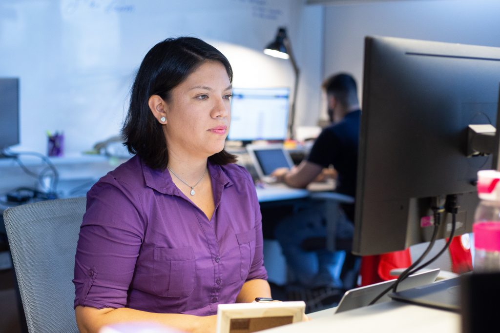 Foto mostra Cecilia Gutierrez sentada em frente a um computador. Ela está usando uma camisa roxa.