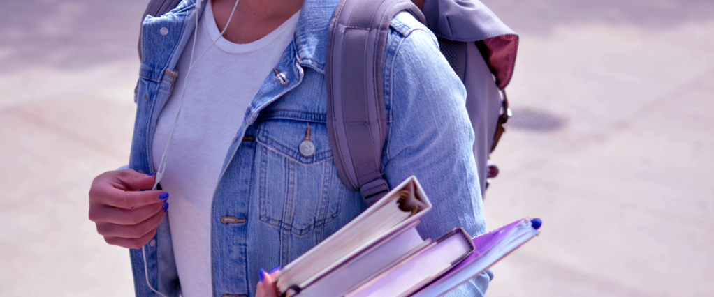 foto mostra tronco de uma jovem de camiseta branca, jaqueta jeans e mochila, carregando livros