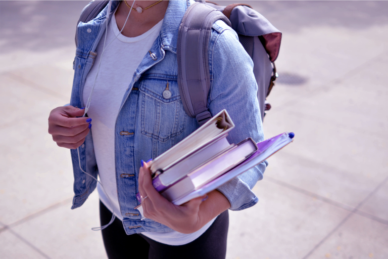 foto mostra tronco de uma jovem de camiseta branca, jaqueta jeans e mochila, carregando livros