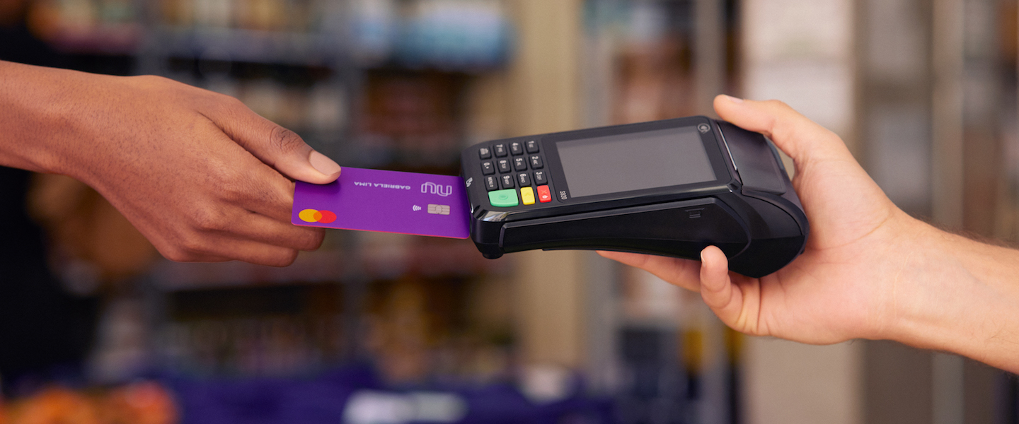 Cartão Nubank Internacional: na foto dentro de uma loja, uma mão segura uma máquina preta de cartão enquanto outra insere o cartão Nubank nela.