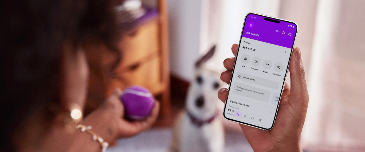 Como declarar a conta do Nubank no IR- Na imagem, uma mulher negra segura um celular com a aplicativo do Nubank na mão direita e uma bolinha roxa na mão esquerda. Ao fundo, um cachorro branco olha para a bolinha na mão da mulher.