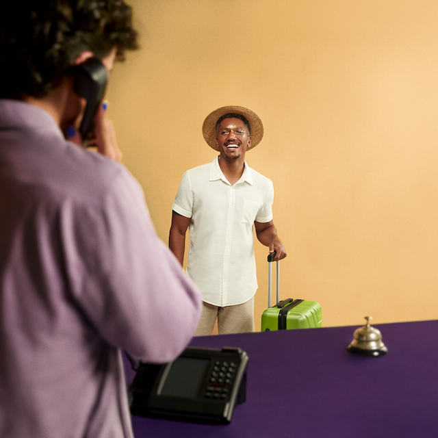 Imagem de um homem negro com uma mala de viagem, em frente ao balcão de um hotel. Ele veste camisa branca, chapéu de palha e sorri. Ele está com o rosto pintado com glitter dourado.