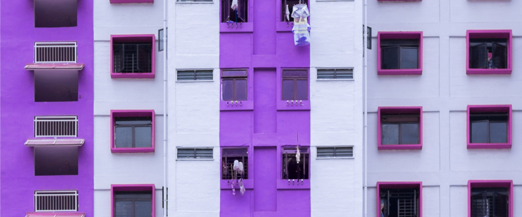 Imagem mostra a fachada de um prédio com muitas janelas. A faixa central do prédio e as laterais são roxas. Os beirais das demais janelas são rosa escuro