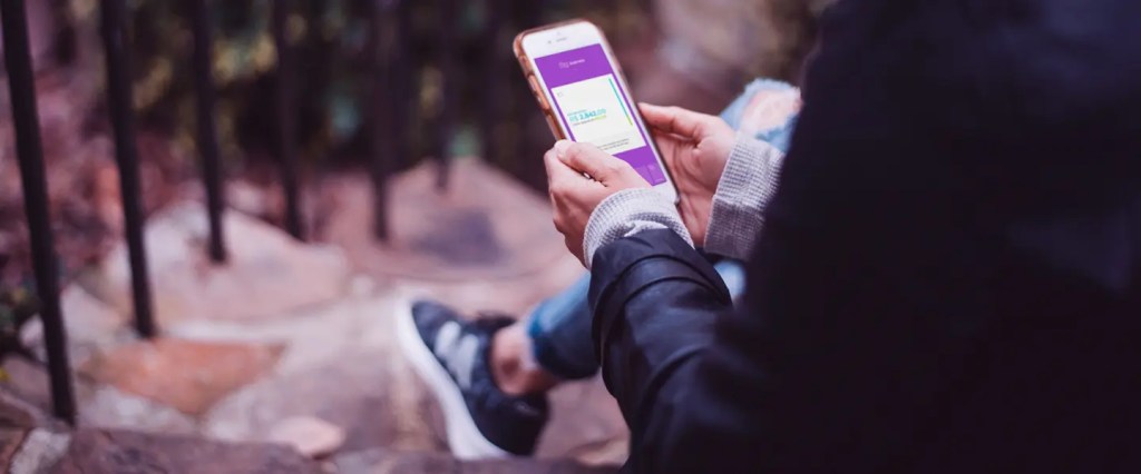 NuConta: Pessoa sentada em uma escada ao ar livre segura um telefone com as duas mãos. Na tela, o app do Nubank aberto na aba de NuConta