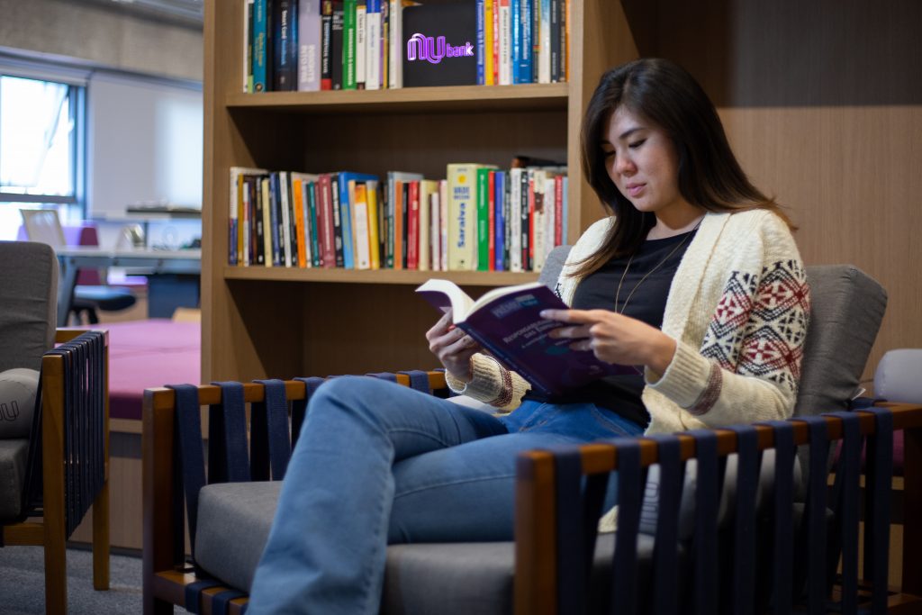 Jovem sentada em uma poltrona na biblioteca do Nubank. Atrás dela, uma parede de livros.