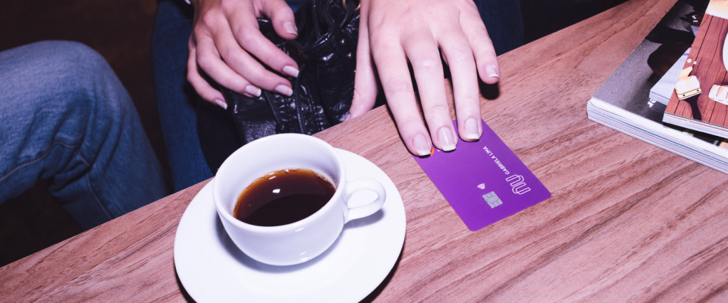 Mesa com uma xícara de café sobre um pires e um cartão de crédito Nubank. Uma mão está segurando o cartão