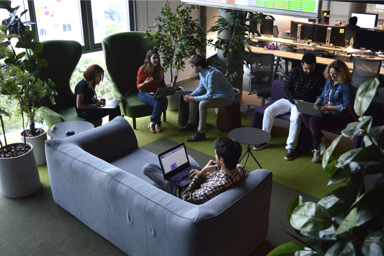 Pessoas sentadas em poltronas e sofás, conversando, em uma área do escritório do Nubank repleta de plantas