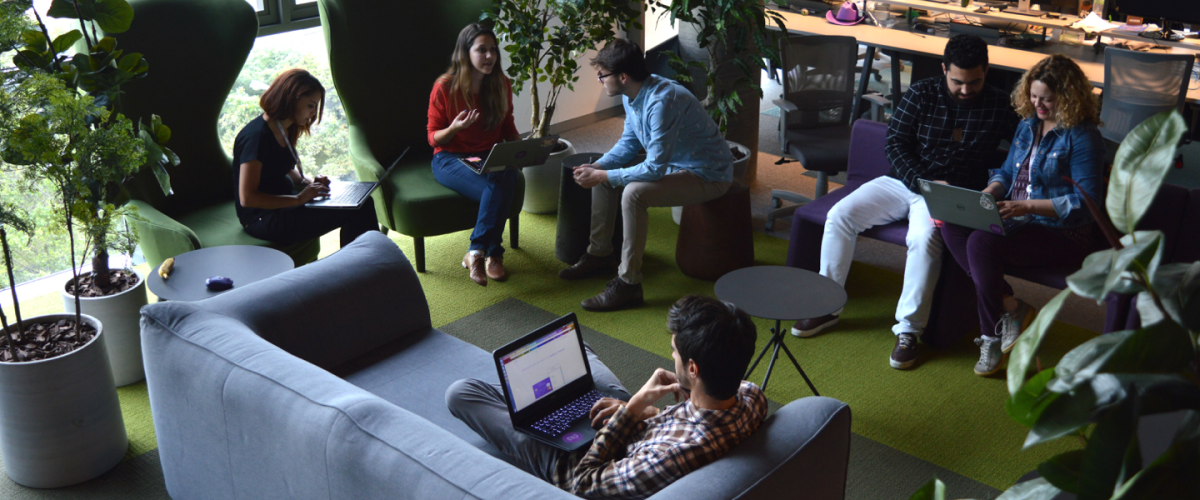 Pessoas sentadas em poltronas e sofás, conversando, em uma área do escritório do Nubank repleta de plantas