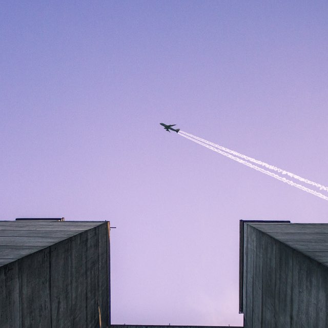 Foto de um avião passando em cima de dois prédios, deixando um rastro de fumaça no céu arroxeado