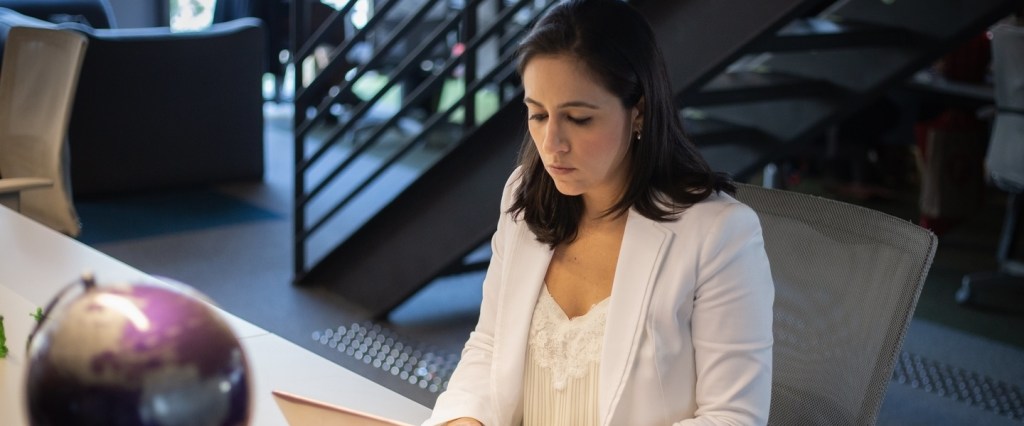 Nossa cofundadora, Cristina Junqueira, vestida de blazer branco e sentada em uma cadeira mexendo em seu notebook.