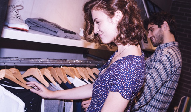 Uma jovem de vestido roxo e cabelos curtos olha para uma arara de roupas em uma loja. Ao fundo, um rapaz também mexe nas camisetas da arara.