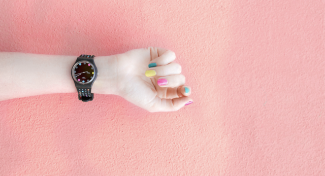 Braço estendido sobre um fundo rosa claro. As unhas das mãos estão pintadas cada uma de uma cor diferentes e, no pulso, há um relógio preto