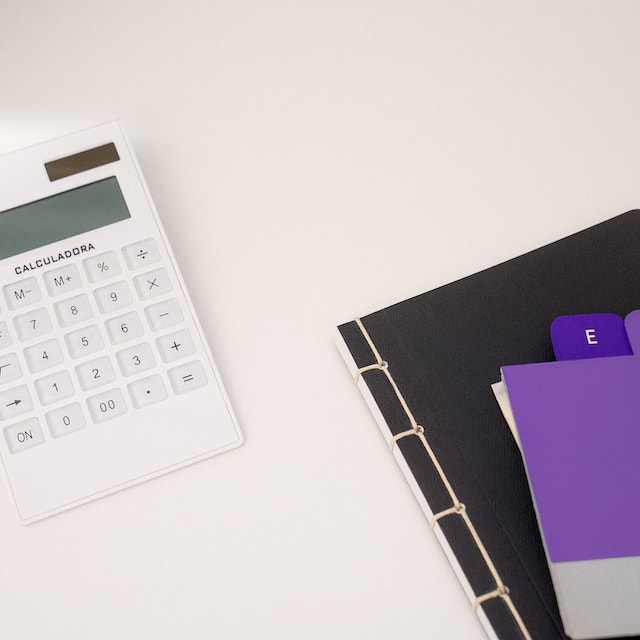 Imagem de uma calculadora branca em cima de uma superfície clara, ao lado uma pasta preta com folhas em tom roxo por cima.