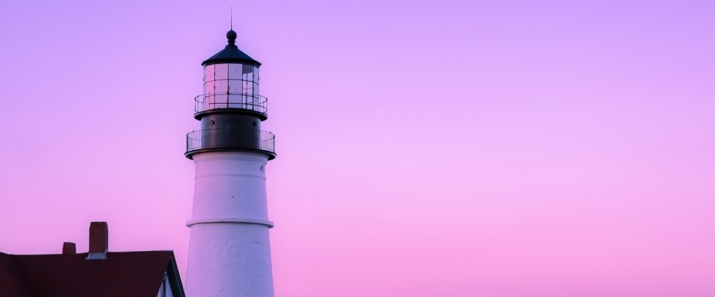 Para onde viajar: um farol à beira mar e o céu em tons de rosa e roxo.