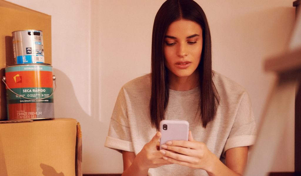 Foto de uma mulher sentada com um celular na mão. Ela tem cabelo liso e comprido e está usando uma camiseta cinza.