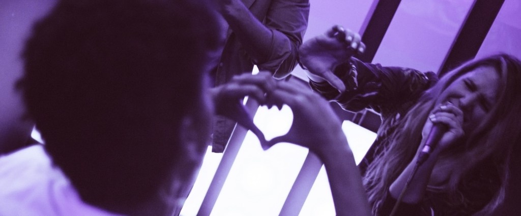 imagem com filtro roxo de um fã fazendo um coração com as mãos para uma cantora no palco