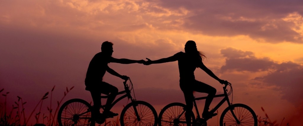 Seguro viagem: duas pessoas seguram mãos enquanto andam de bicicleta ao entardecer