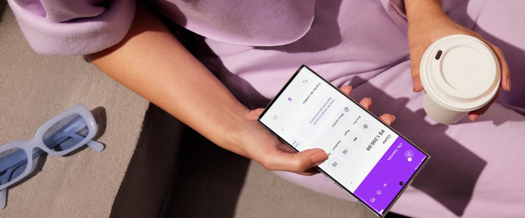 Seu dinheiro está seguro no Nubank? Imagem de uma pessoa segurando um celular com a tela inicial do app do Nubank na mão direita. Na mão esquerda, um copo de café para viagem. A pessoa tem pele branca e veste uma roupa na cor roxa. Na parte esquerda inferior da imagem, um óculos de sol com armação azul celeste sobre a mesa ao lado da pessoa.