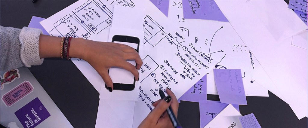 Desenvolvimento de produto Nubank: mesa com pedaços de papel branco e roxo e mãos mexendo em um celular e escrevendo em uma das folhas de papel