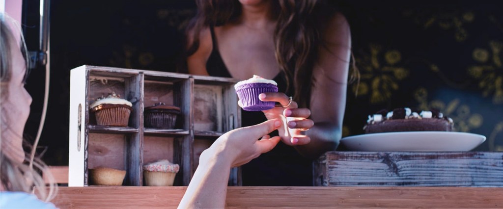 Conta PJ Nubank: imagem mostra uma jovem vendendo um cupcake roxo e uma mão estendida para pegar o produto