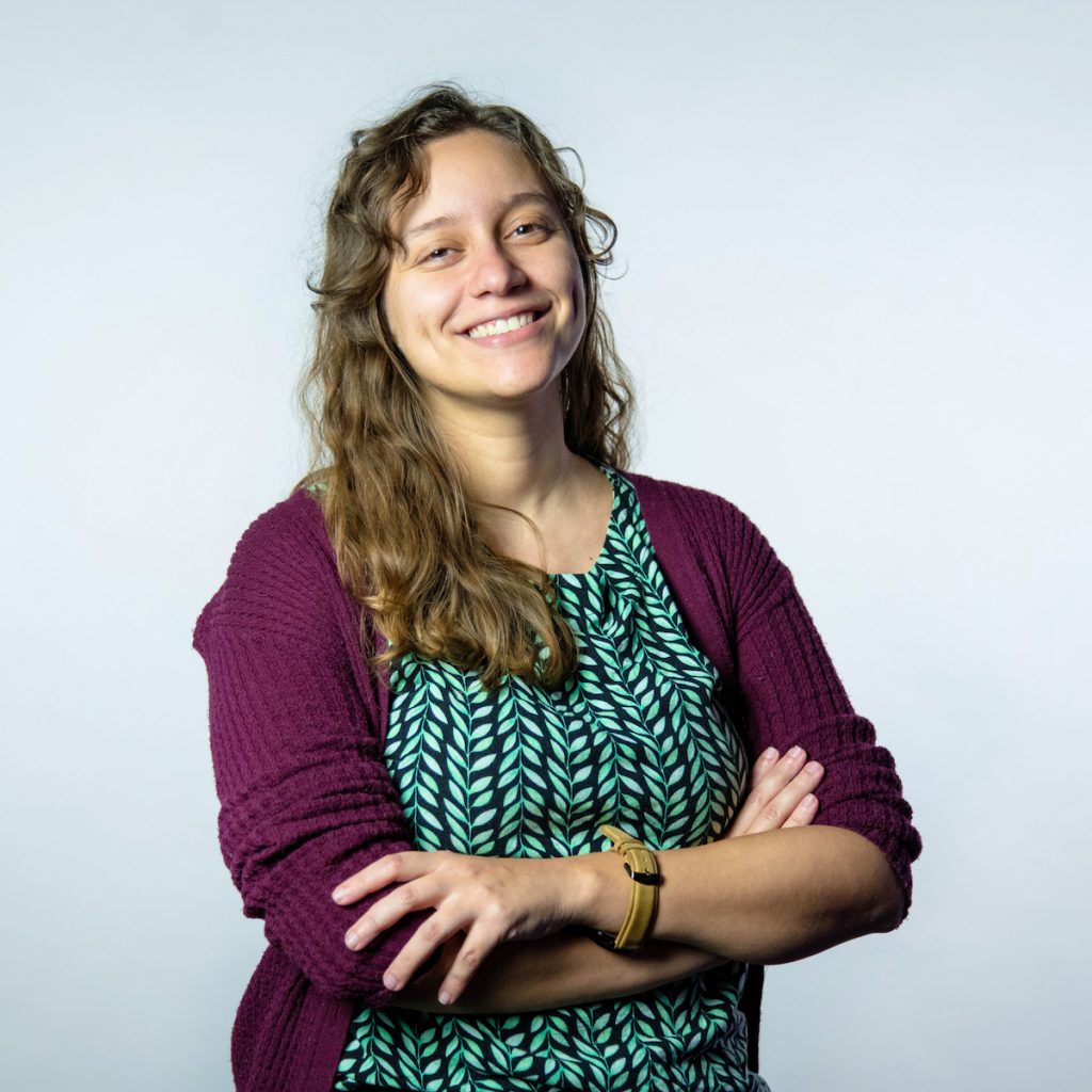 Foto da Jullie Utsch, engenheira de software do Nubank. Ela veste um cardigan vinho, uma blusa verde e está com os braços cruzados. Ela é loira, tem os cabelos ondulados, está sorrindo e tem covinhas na bochecha.
