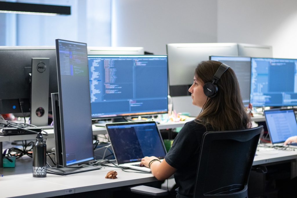 Uma mulher trabalha com códigos de programação em três telas de computador.