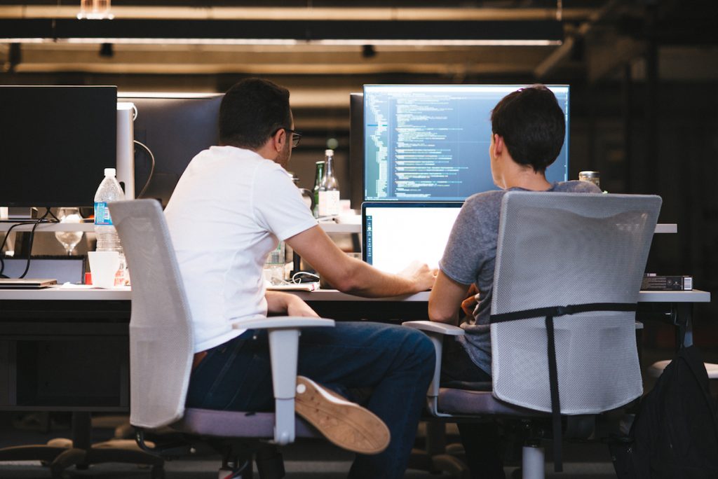 Dois homens sentados, trabalhando juntos num computador e um código de programação na tela.