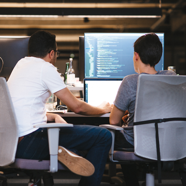 Engenharia de software Nubank: dois homens sentados olhando para um computador. Na segunda tela, um programa de código de programação.