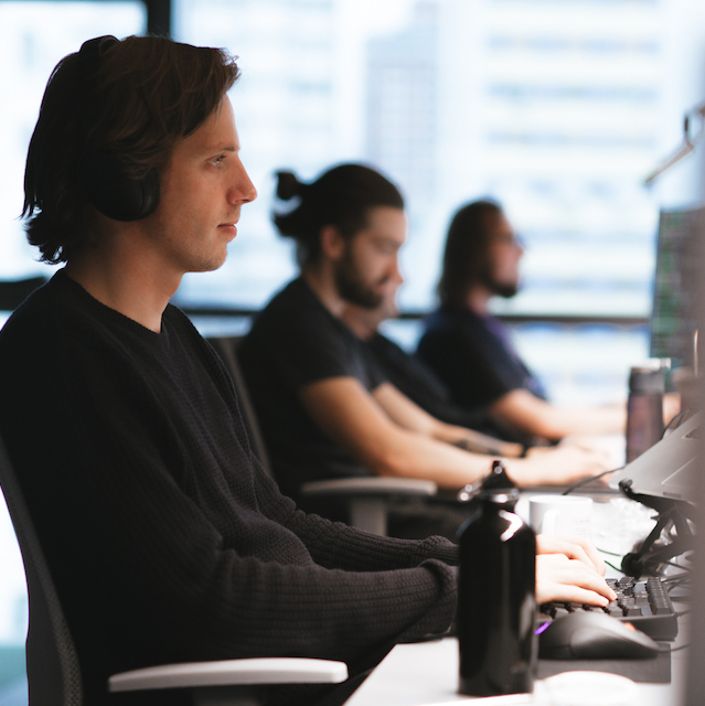 Machine learning: foto de um homem trabalhando. Ele veste preto, usa fones de ouvido e está digitando no teclado. Mais ao fundo, outros dois homens também estão trabalhando em seus computadores.