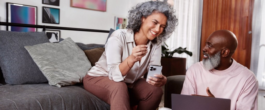 Tipos de empréstimo - imagem de uma mulher branca ao lado de um homem negro, e ambos estão sorrindo. Ela segura um celular com uma das mãos e veste uma camisa de seda off white e uma calça de linho marrom. Ela tem cabelos grisalhos, cacheados, na altura do ombro. O homem veste uma camiseta rosa claro, é careca e tem barba branca e comprida. Eles estão em uma sala de estar, com sofá, almofadas, quadros, plantas e uma mesa de centro com objetos decorativos e um prato com frutinhas.