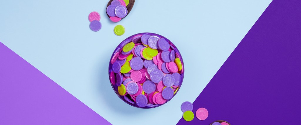 Imagem de um pote cheio de moedas coloridas, com duas colheres ao lado dele. O pote está sobre uma mesa em tons de roxo e azul.