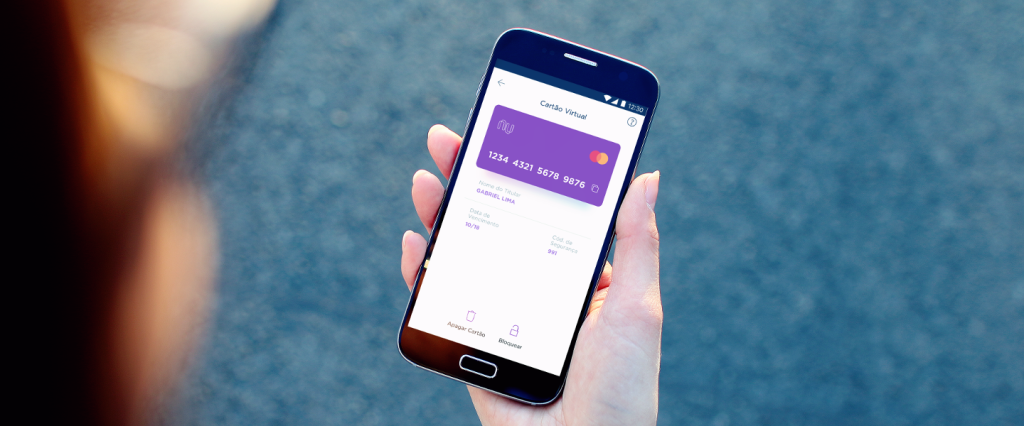 Cartão Virtual Nubank: Mão segura um telefone. Na tela, a imagem de uma cartão de crédito roxo.